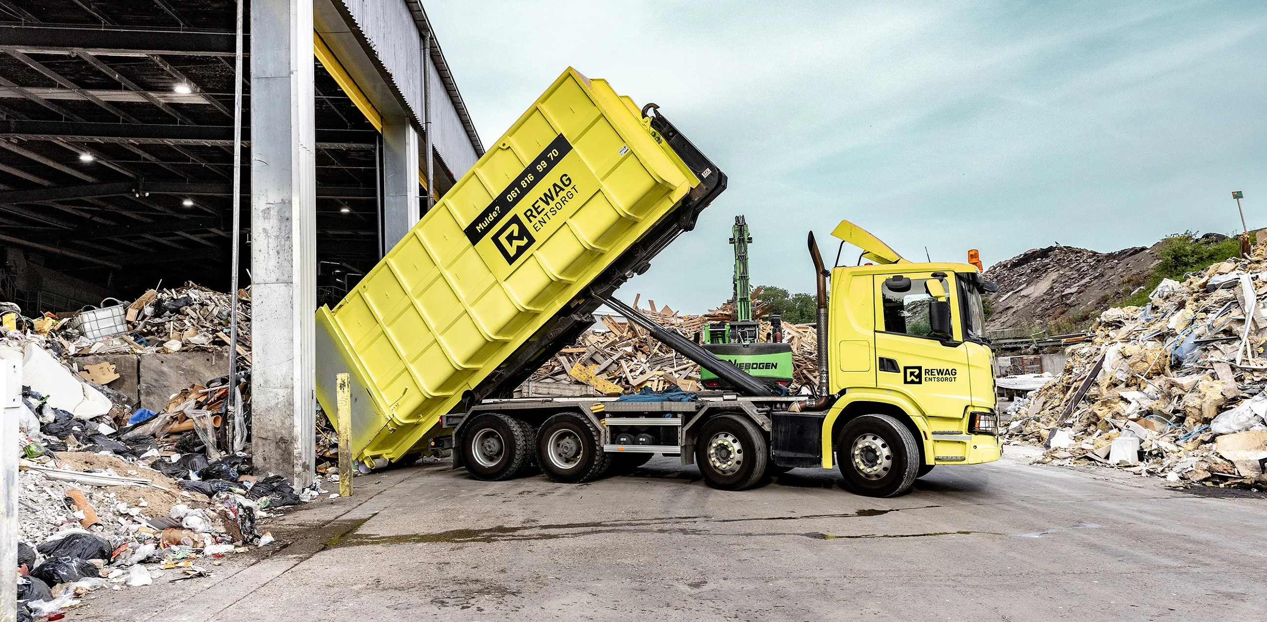 Gelber REWAG-Lastwagen kippt eine Mulde vor der offenen Sortierhalle, dahinter Bagger und Materialhaufen