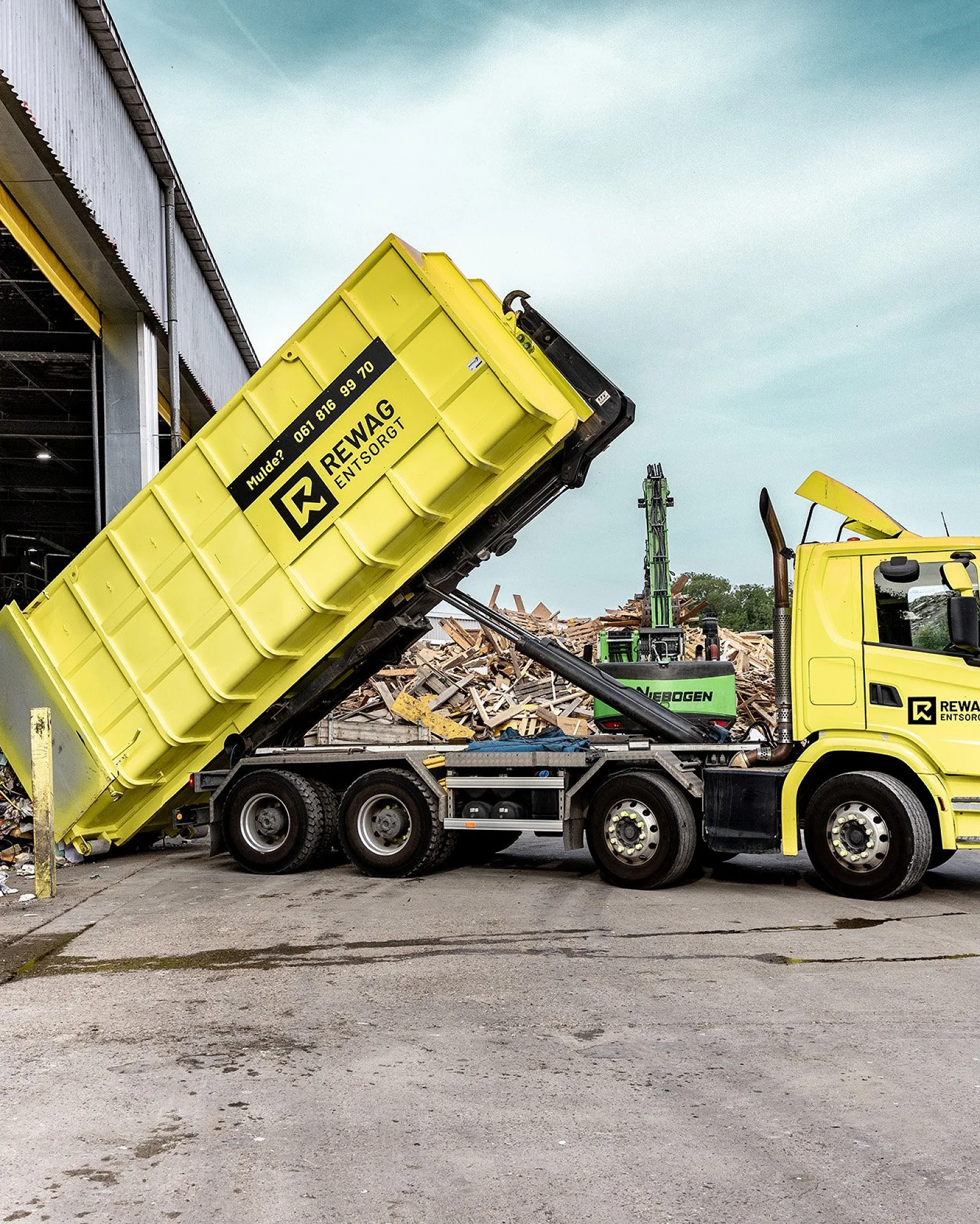 Gelber REWAG-Lastwagen kippt eine Mulde vor der offenen Sortierhalle, dahinter Bagger und Materialhaufen
