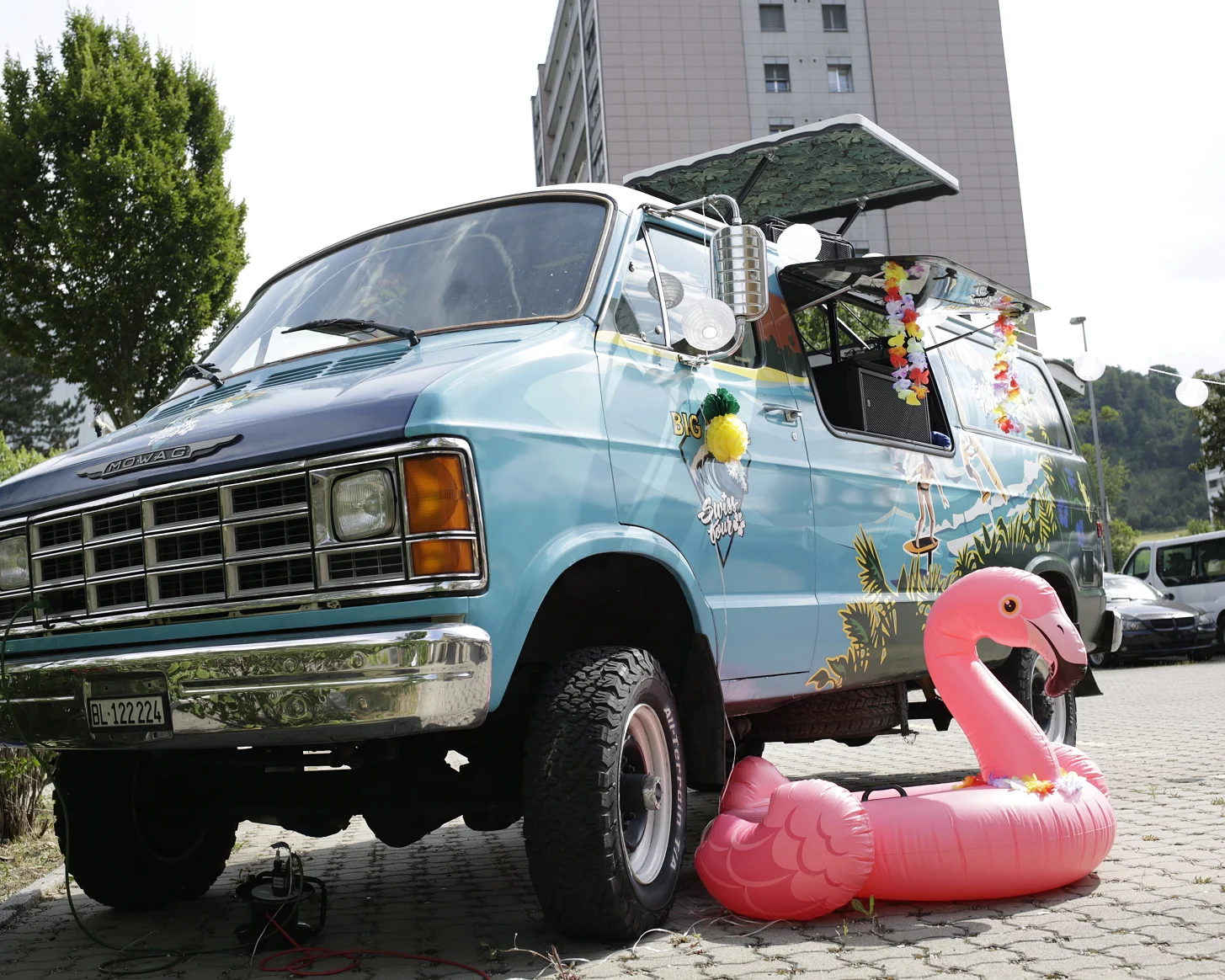 Hellblauer Van mit geöffneter Seitenklappe, davor ein rosa Flamingo-Schwimmring