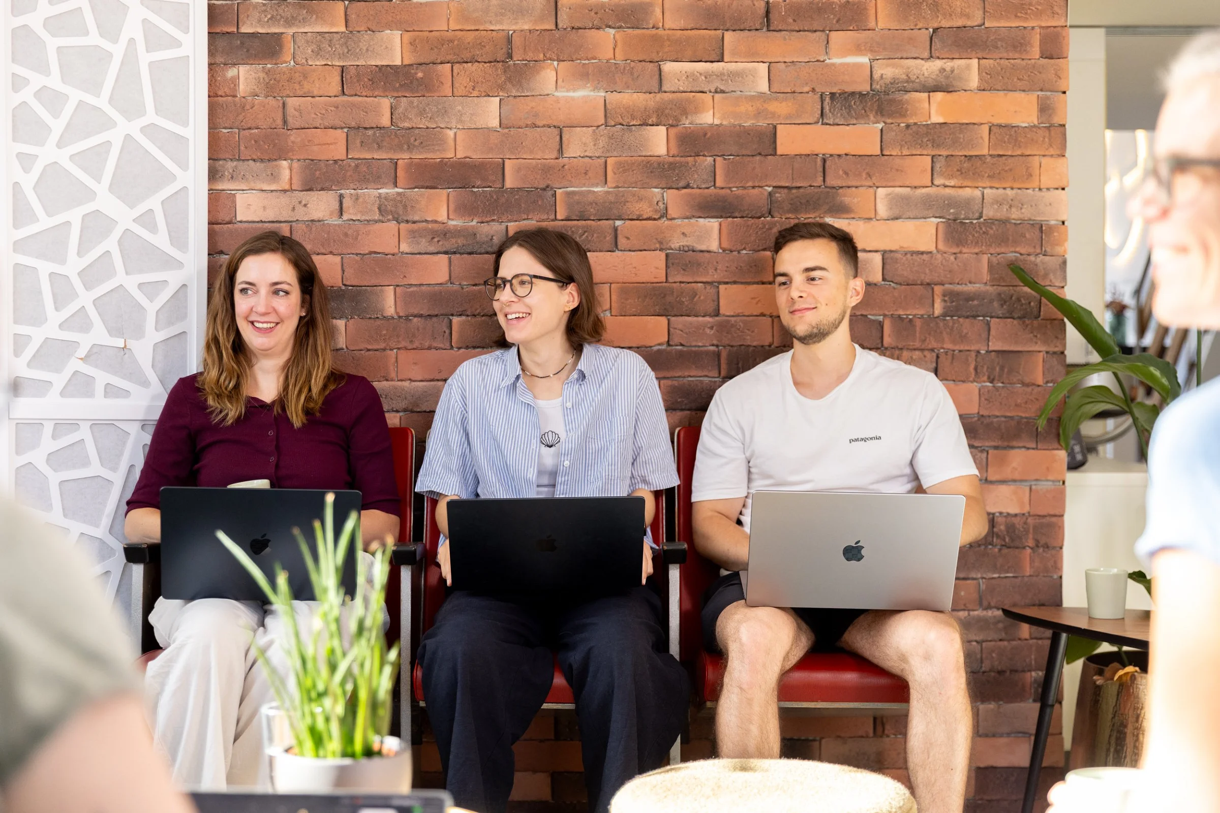 Drei Personen sitzen mit Laptops vor der Backsteinwand im Atelier