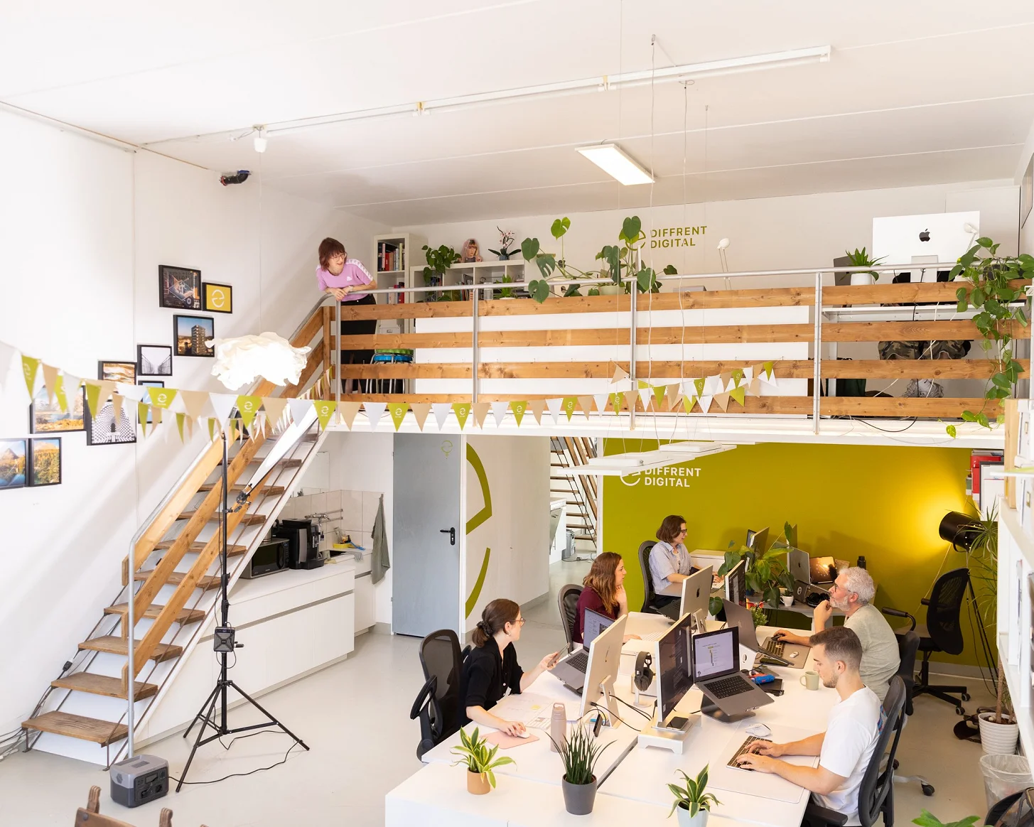 Das Atelier von der Galerie aus: Wimpelkette, Holztreppe und die Arbeitsplätze vor der gelben Wand