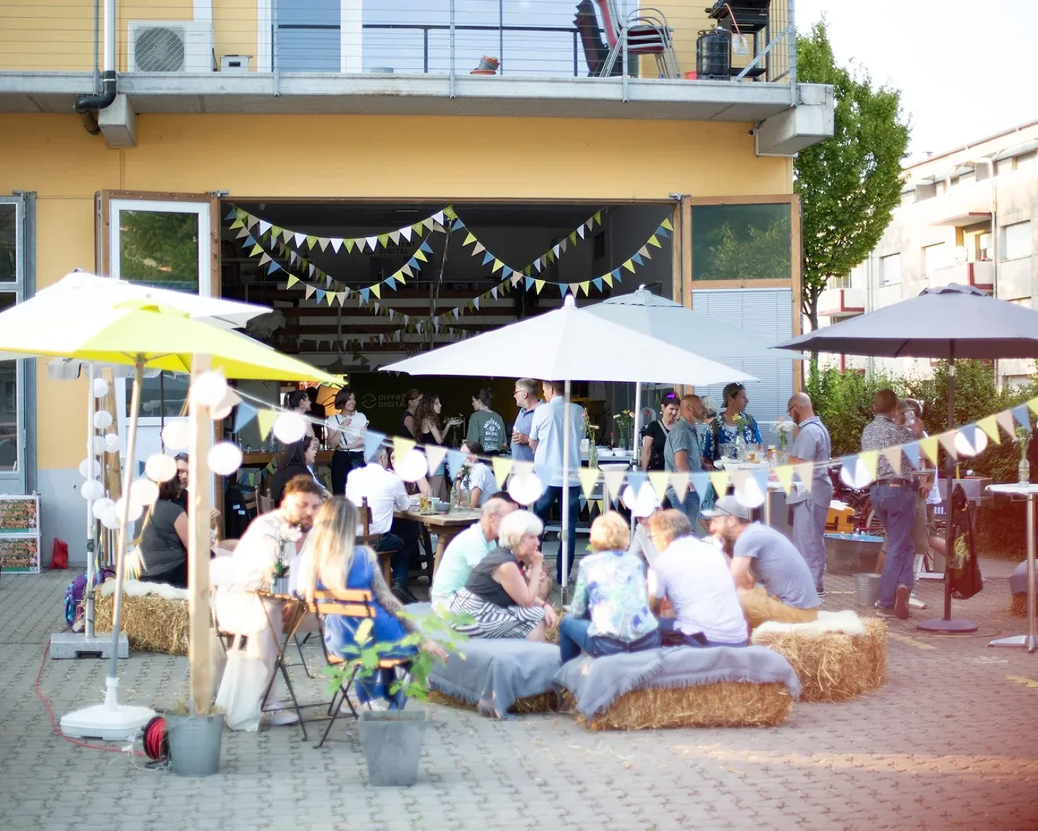 Abendstimmung am Fest: Sonnenschirme, Lichterketten und Gäste auf Strohballen vor dem Büro