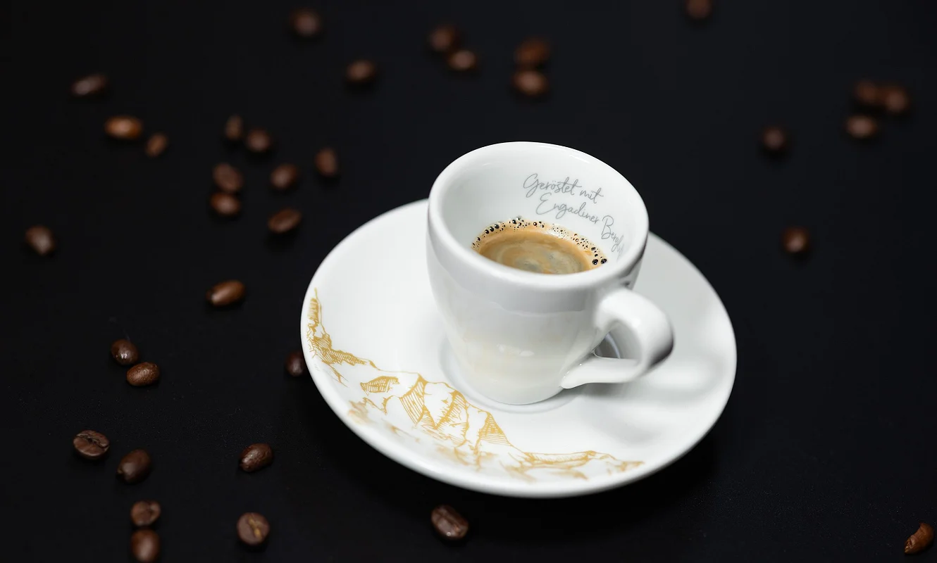 Espressotasse mit Bergillustration auf der Untertasse, drumherum geröstete Kaffeebohnen auf schwarzem Grund