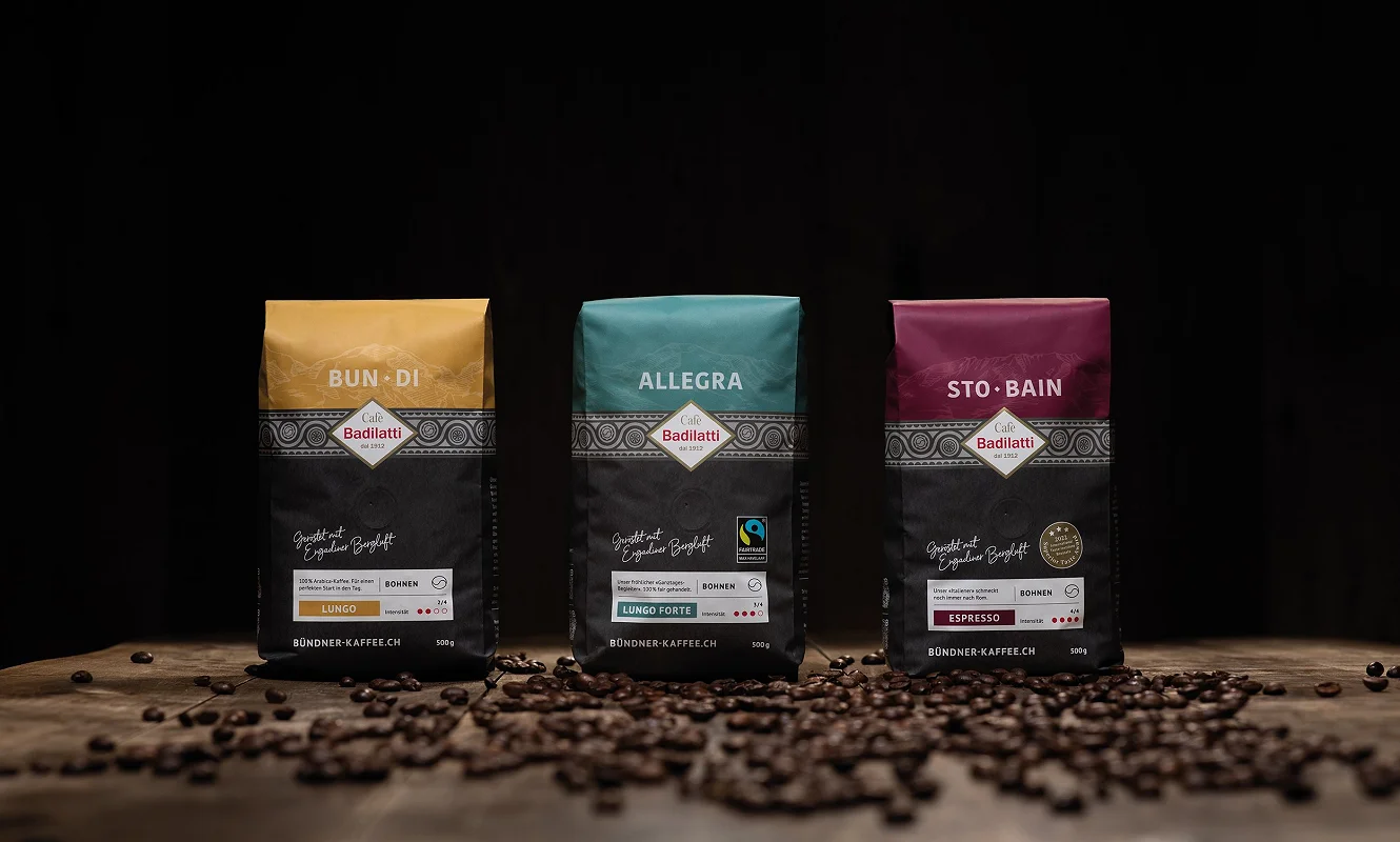 Drei Kaffeepackungen nebeneinander: «Bun·Di», «Allegra» und «Sto·Bain» vor gestreuten Bohnen