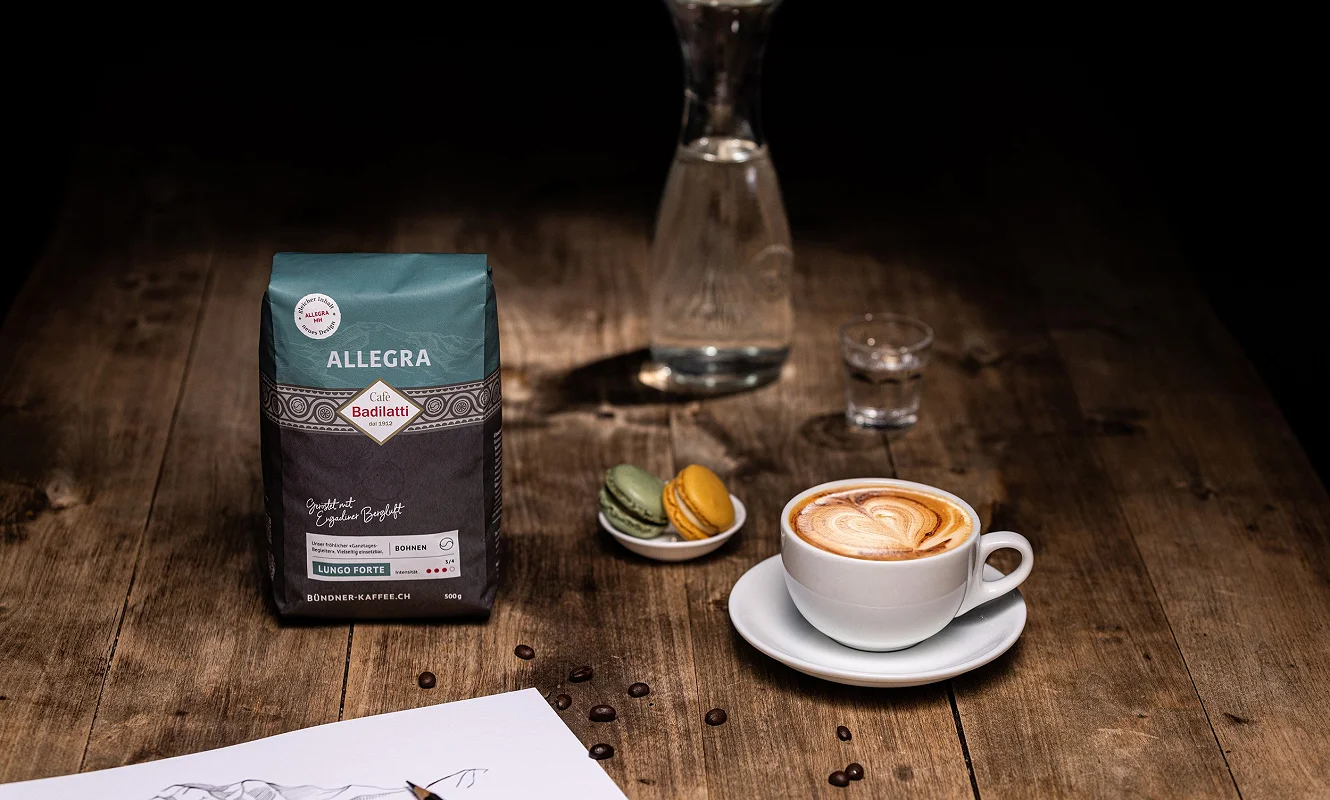 Packung «Allegra» neben Cappuccino, Macarons und einer Bergzeichnung auf dem Holztisch