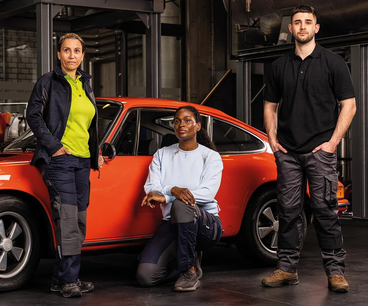 Drei Personen in Arbeitskleidung vor einem roten Oldtimer in der Werkstatt