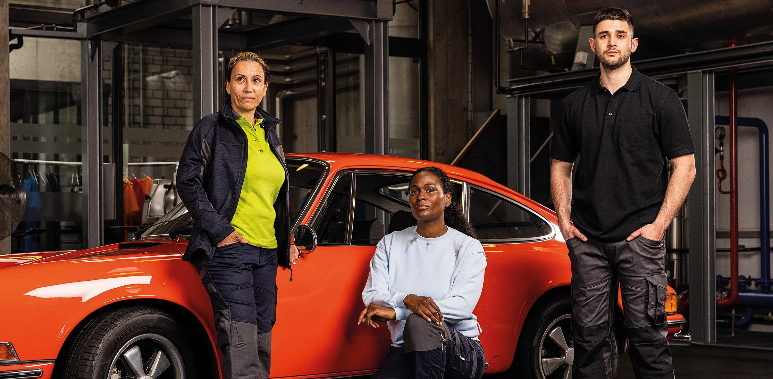 Drei Personen in Arbeitskleidung vor einem roten Oldtimer in der Werkstatt