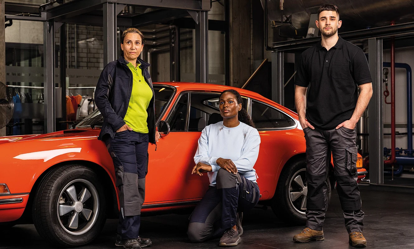 Drei Personen in Arbeitskleidung vor einem roten Oldtimer in der Werkstatt