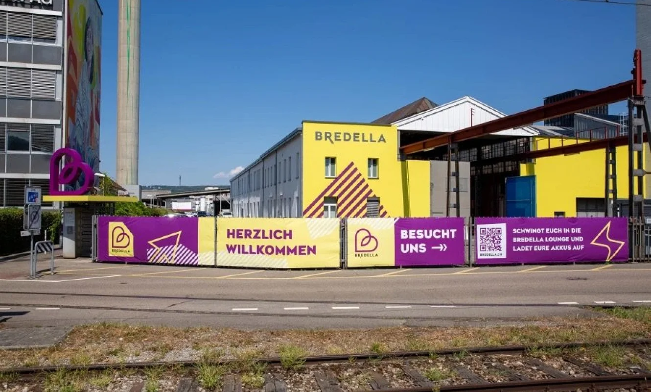 Bauzaun am Areal mit den Bannern «Herzlich willkommen» und «Besucht uns», dahinter die gelbe Bredella-Halle