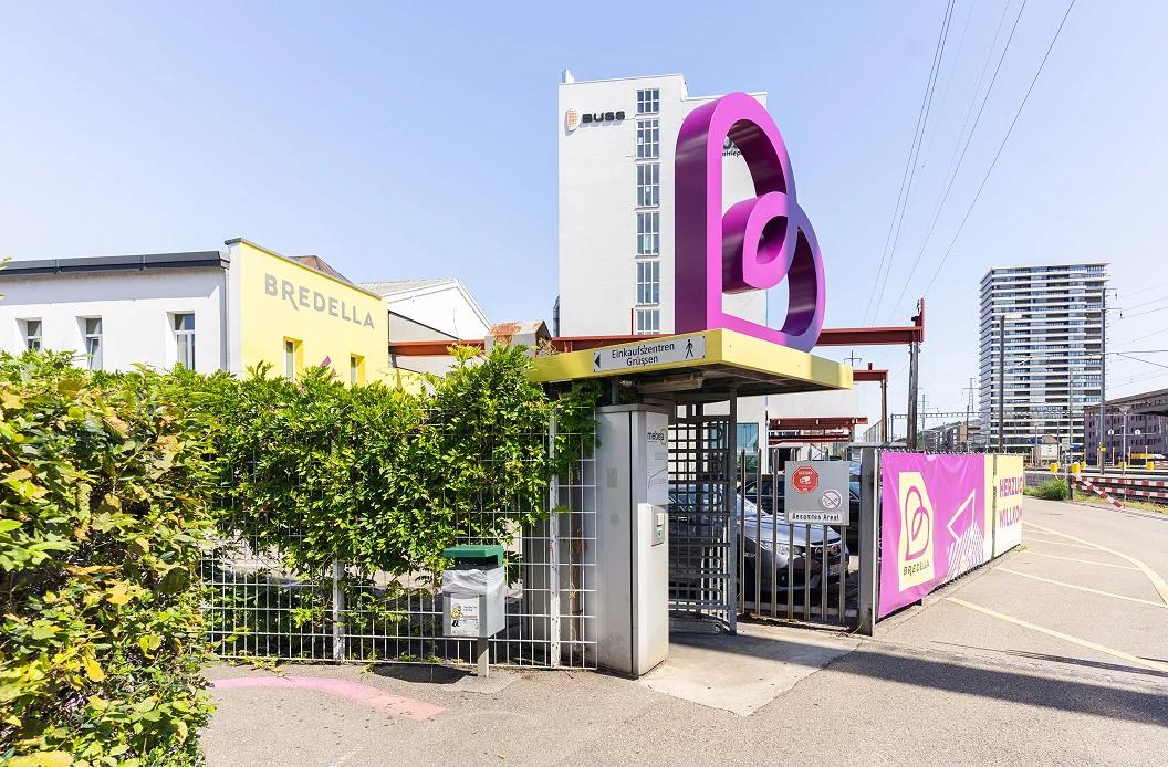 Arealeingang mit dem Bredella-Signet als Skulptur über dem Zugang, dahinter das Buss-Hochhaus