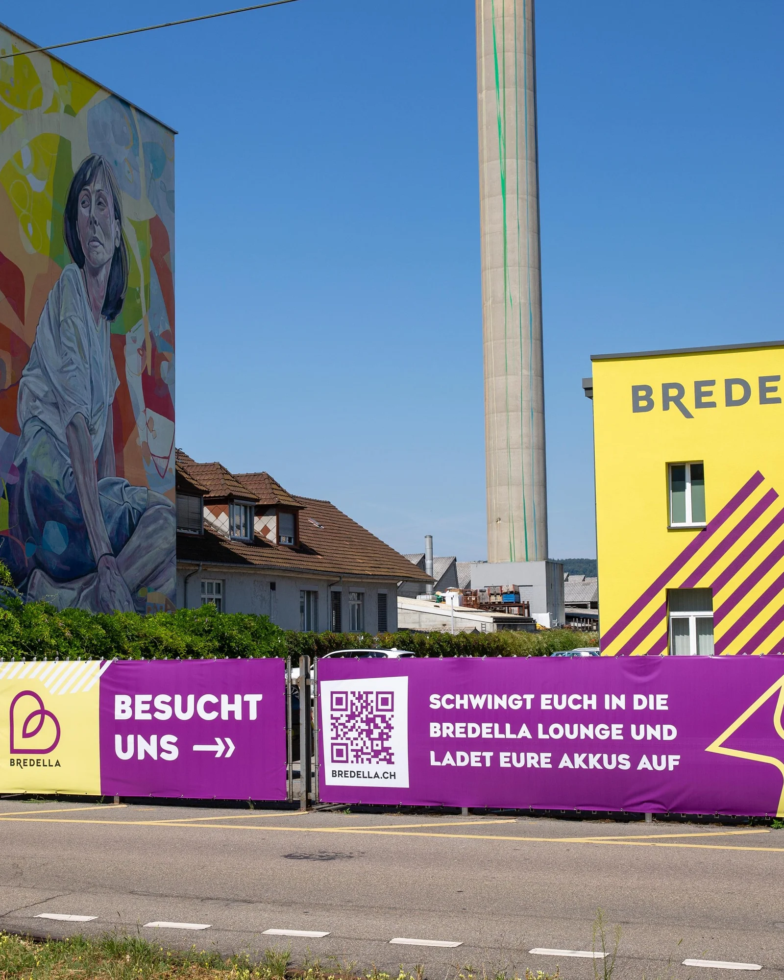 Bauzaun-Banner vor der gelben Bredella-Halle, links das Wandbild an der Rohrbogen AG