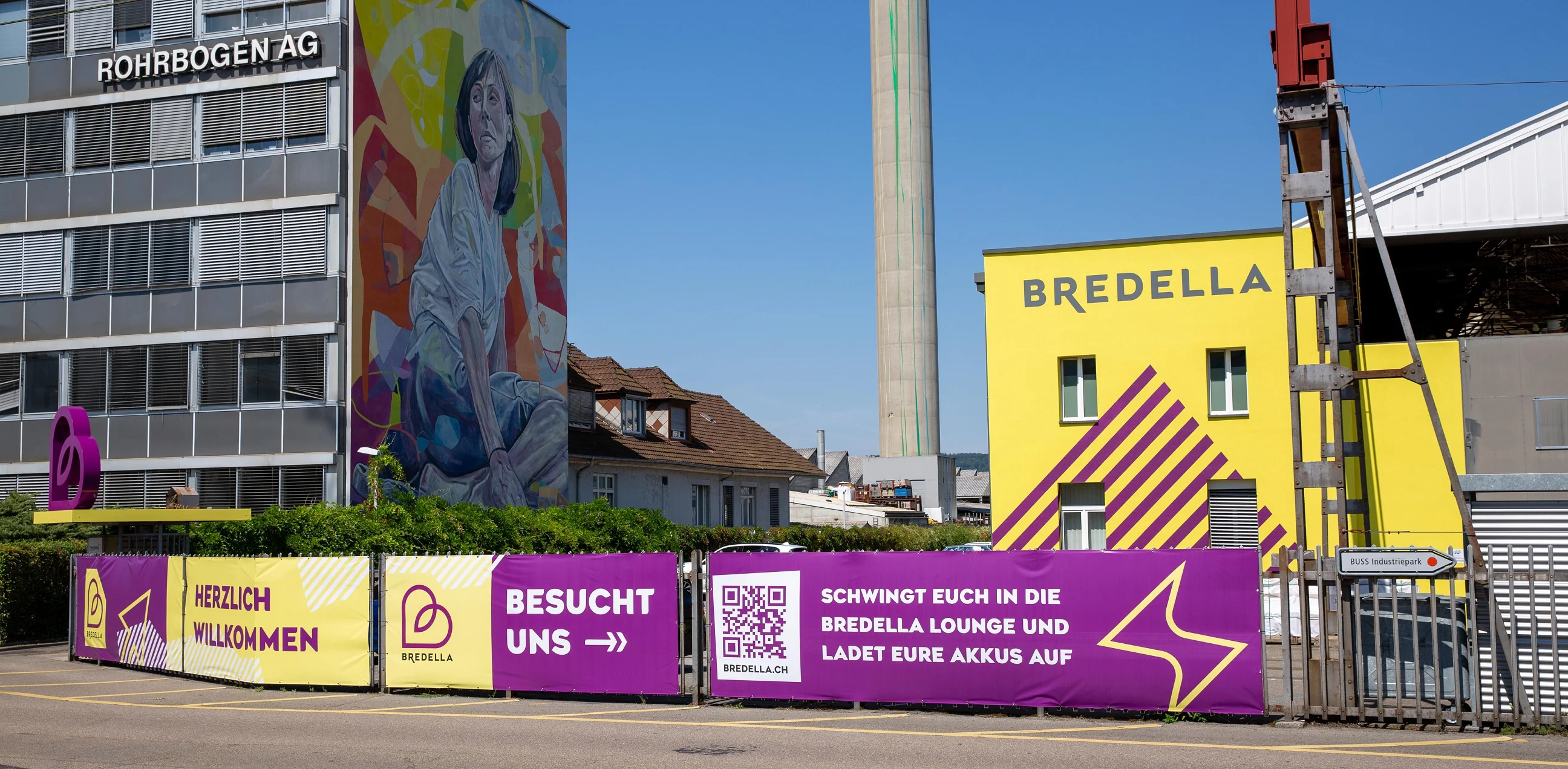 Bauzaun-Banner vor der gelben Bredella-Halle, links das Wandbild an der Rohrbogen AG
