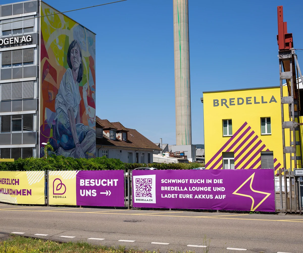 Bauzaun-Banner vor der gelben Bredella-Halle, links das Wandbild an der Rohrbogen AG