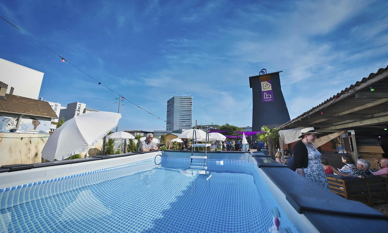 Pool im Bredella Village mit Sonnenschirmen, dahinter der Salzbohrturm mit dem Village-Banner. Foto Marc Gilgen