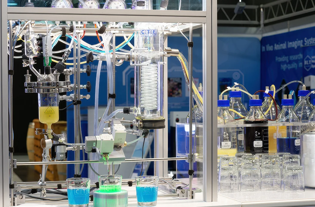 Die Cocktailmaschine am CTE-Messestand aus der Nähe: Laboranlage aus Glas und Aluminium mit Reaktor, Kühler, Ventilen und Schläuchen, davor drei Gläser mit blauem und grünem Getränk