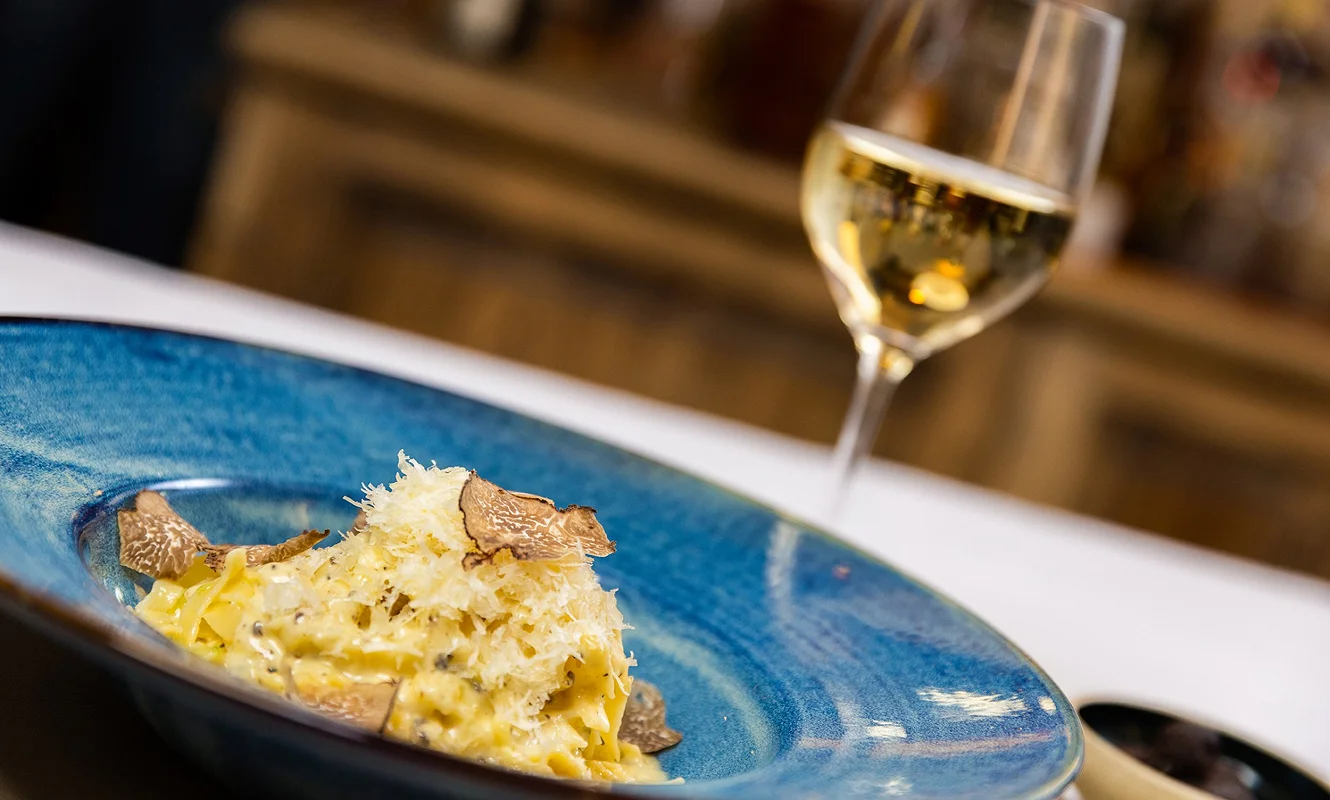 Tagliatelle mit Trüffel und Hartkäse im blauen Teller, dahinter ein Glas Weißwein