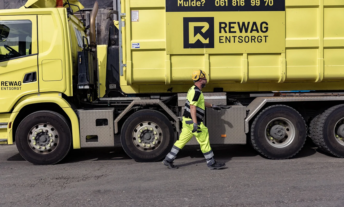 Mitarbeiter in Warnkleidung geht am gelben Lastwagen entlang, auf der Mulde steht «Mulde? 061 816 99 70»