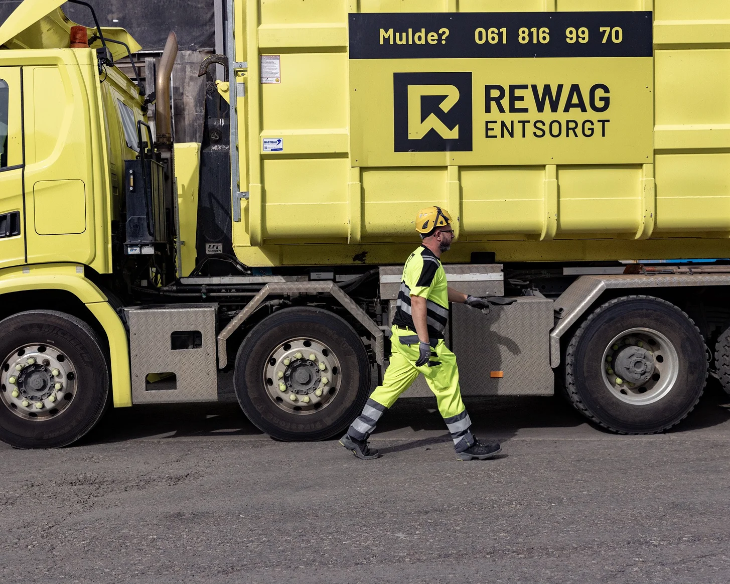 Mitarbeiter in Warnkleidung geht am gelben Lastwagen entlang, auf der Mulde steht «Mulde? 061 816 99 70»