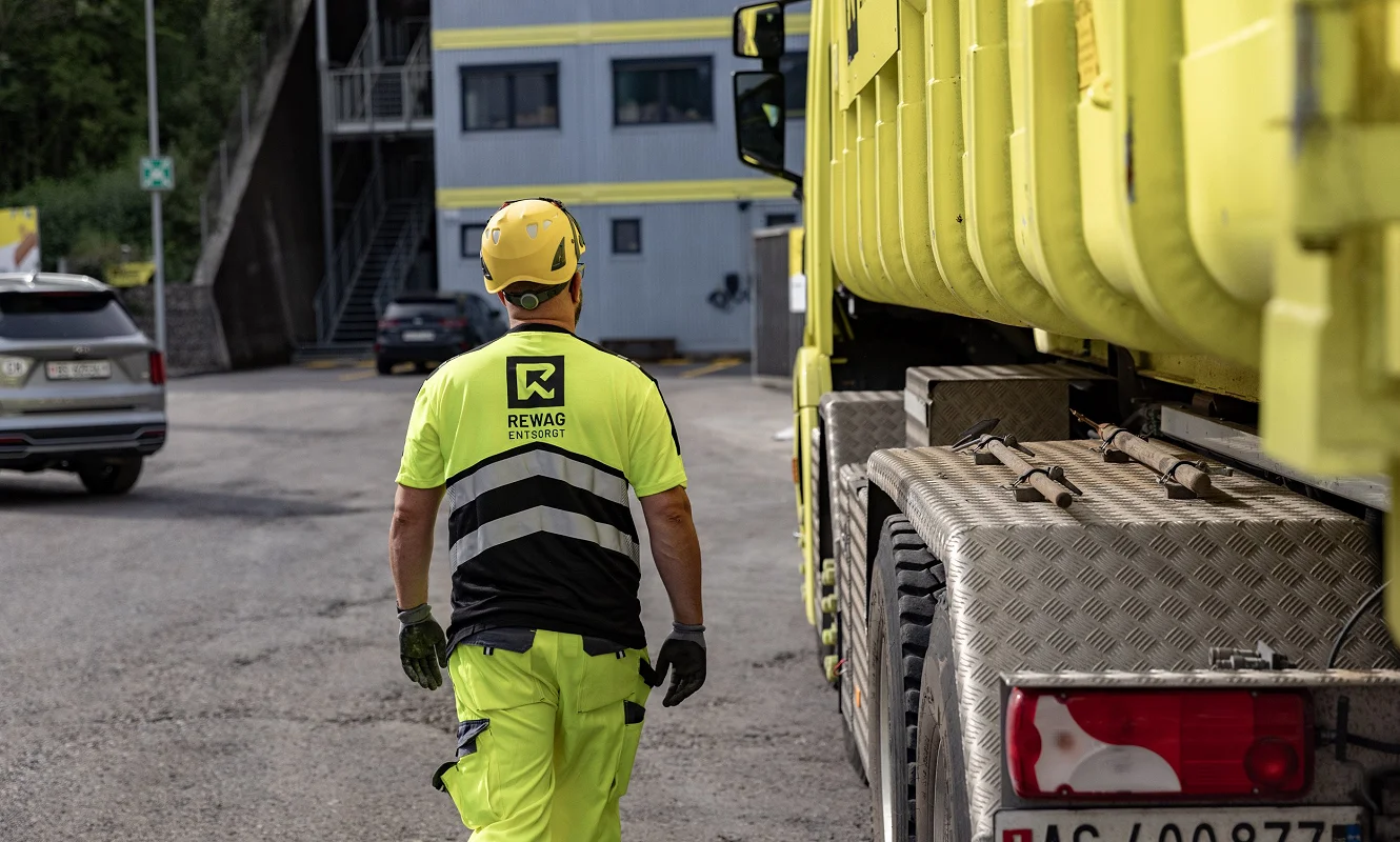 Mitarbeiter mit Helm und Warnkleidung von hinten neben dem Lastwagen auf dem Areal