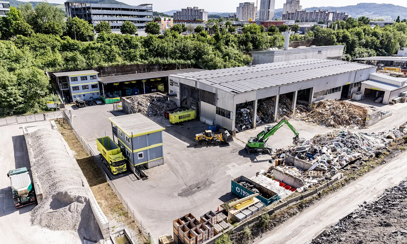 Luftbild des REWAG-Areals mit Sortierhalle, Mulden und Materialhaufen, dahinter die Stadt