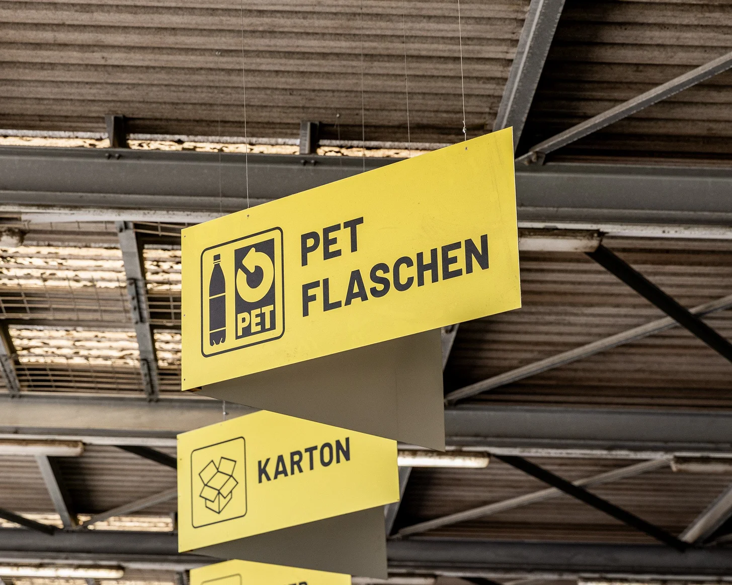Abgehängte Schilder in der Halle: «PET Flaschen» und «Karton» mit Piktogrammen