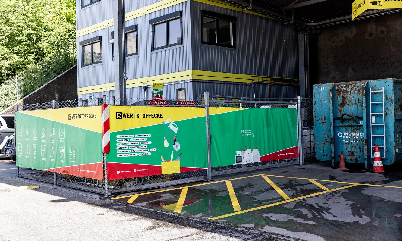 Umzäunte «Wertstoffecke» mit grün-roter Banderole und Piktogrammen der zulässigen Materialien