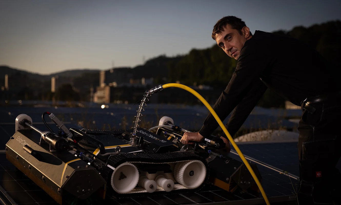 Reinigungsroboter auf einer Photovoltaikanlage, daneben eine Person mit Wasserschlauch in der Dämmerung