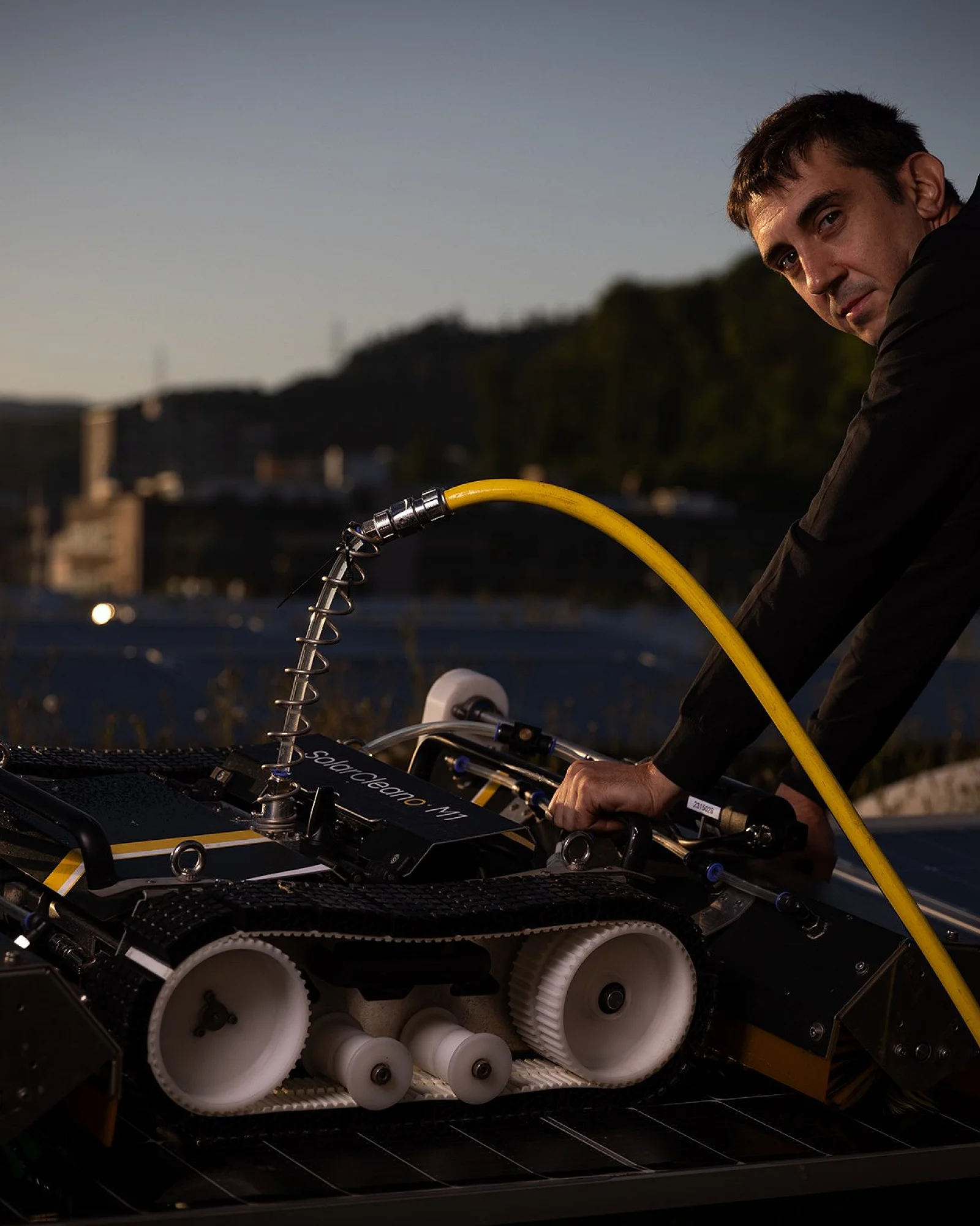 Reinigungsroboter auf einer Photovoltaikanlage, daneben eine Person mit Wasserschlauch in der Dämmerung
