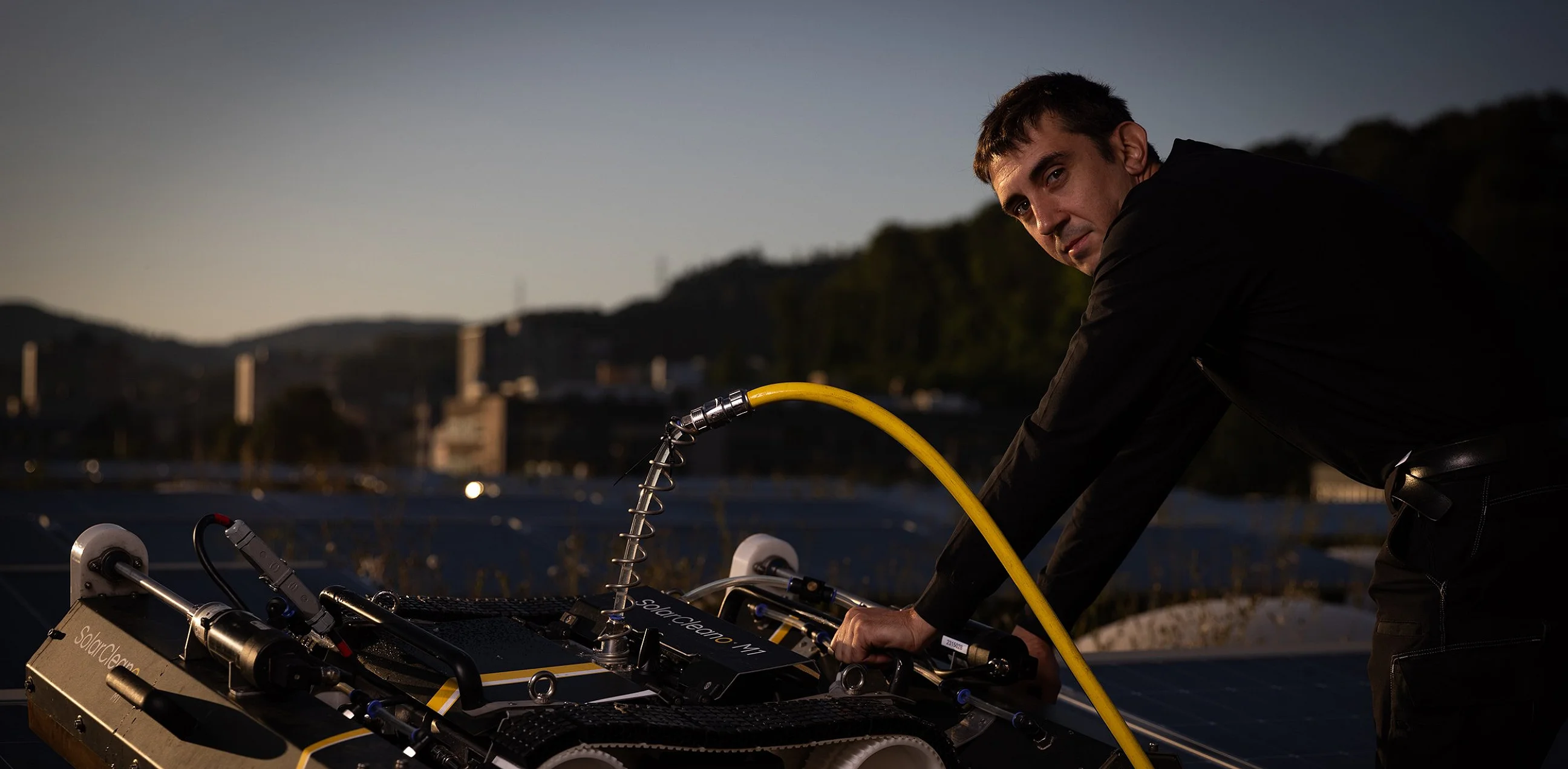 Reinigungsroboter auf einer Photovoltaikanlage, daneben eine Person mit Wasserschlauch in der Dämmerung
