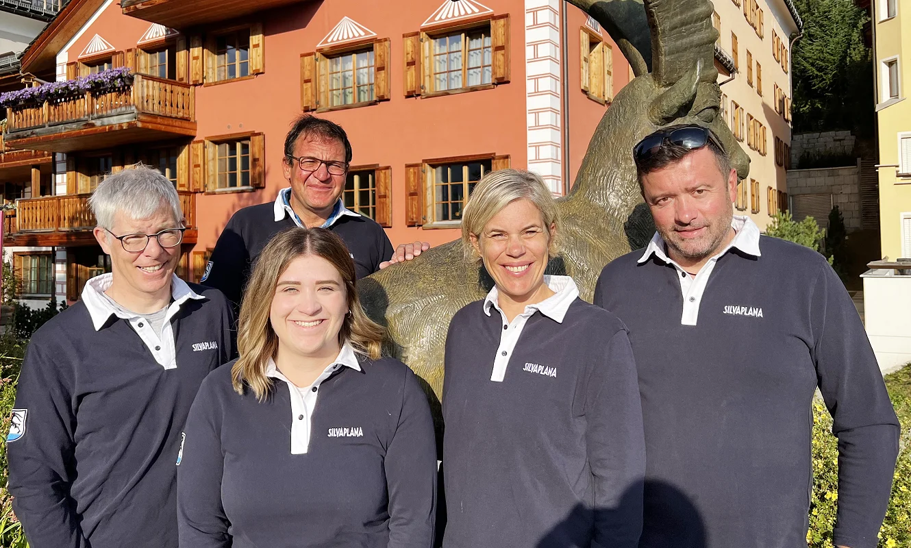 Fünf Personen in Silvaplana-Poloshirts vor einem orangen Engadinerhaus mit Steinbockfigur